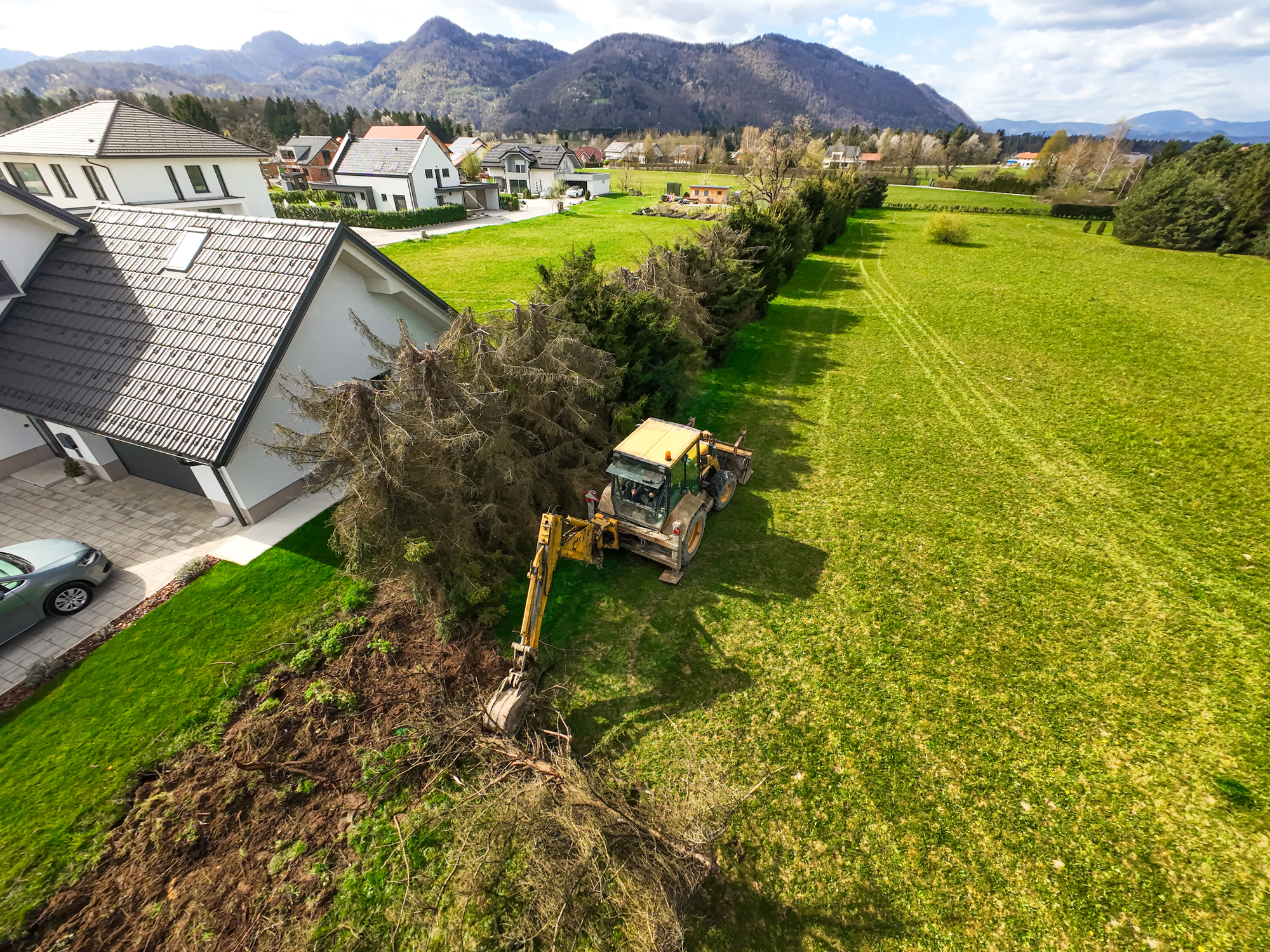 From above, a backhoe loader is seen methodically clearing pine trees from a forested area, its articulated arm uprooting trunks with mechanical precision. The once-dense canopy now shows patches of exposed earth and scattered logs, revealing the transformation brought on by land-clearing operations. The machine's yellow frame contrasts starkly against the deep green of the surrounding forest, emphasizing the intervention of human technology in a natural landscape. This aerial view captures the raw process of forestry work — the intersection of utility, industry, and environment.